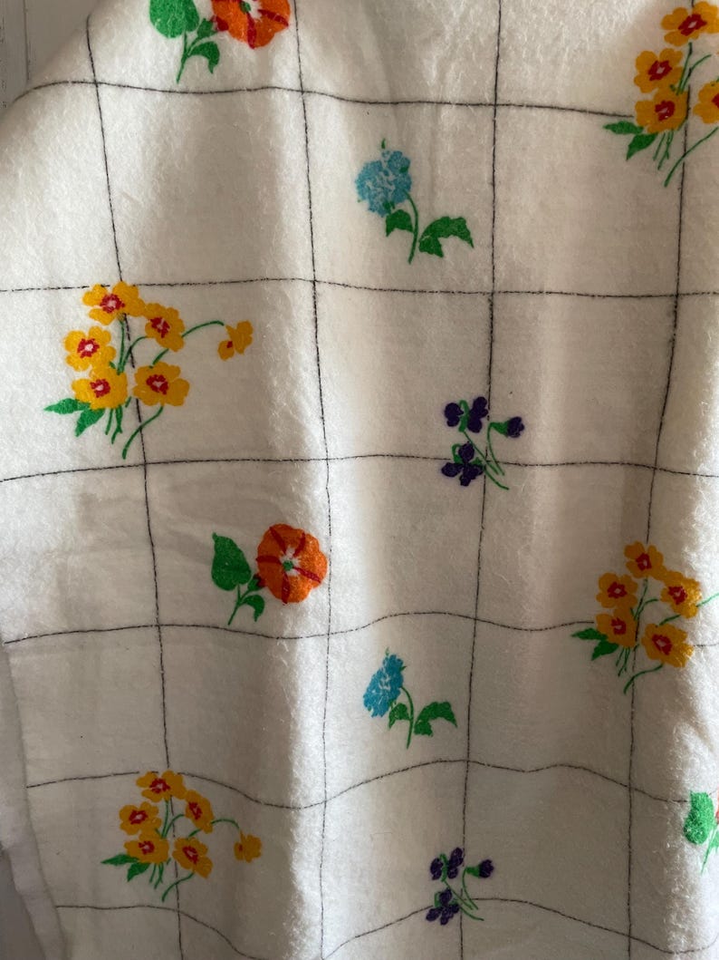 Op de afbeelding: Witte stof met een ruitpatroon, geborduurd met kleurrijke bloemen. De bloemen zijn in tinten van oranje, geel, blauw en paars, met groene bladeren. De stof lijkt zacht en getextureerd.
