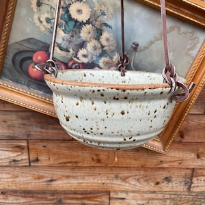 Peut inclure: Un pot de fleurs suspendu en céramique mouchetée avec une sangle en cuir marron. Le pot blanc cassé présente des mouchetures marron et un bord marron. La sangle en cuir est fixée au pot avec des anneaux métalliques et des nœuds. L'arrière-plan comprend une peinture encadrée.