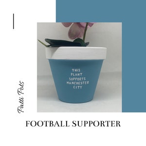 May include: A light blue ceramic planter with white trim. The planter has the text "This plant supports Manchester City" written on it. The planter is decorated with a pink flower and green leaves.