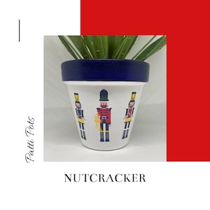 May include: White ceramic planter with a blue rim, featuring a Nutcracker design with three nutcrackers playing instruments. The planter is filled with a green succulent plant.