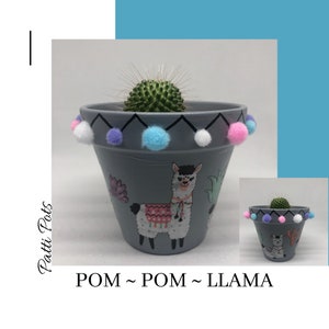 May include: A grey terracotta planter with a painted llama design and a cactus plant inside. The planter is decorated with a row of colourful pom-poms around the rim. The text "POM ~ POM ~ LLAMA" is written below the planter.