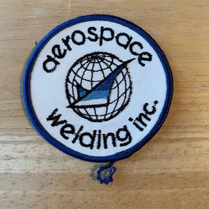 May include: Circular patch with a white centre and a blue border. The patch features the words "aerospace welding inc." in black, surrounding a globe graphic with a blue arrow. It is likely an emblem or logo.