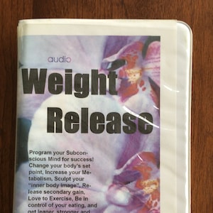 May include: A white cassette tape case with a purple orchid on the front. The text "audio Weight Release" is printed in black on the front of the case. The text below the title reads "Program your Subconscious Mind for success! Change your body's set point, Increase your Metabolism, Sculpt your "inner body image", Release secondary gain, Love to Exercise, Be in control of your eating, and get leaner, stronger and happier with Hypnotherapy."