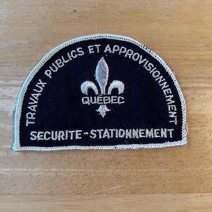 May include: A navy blue patch with white embroidered text and a silver fleur-de-lis emblem. The text reads "TRAVAUX PUBLICS ET APPROVISIONNEMENT" and "SÉCURITÉ - STATIONNEMENT" with "QUÉBEC" below the emblem.