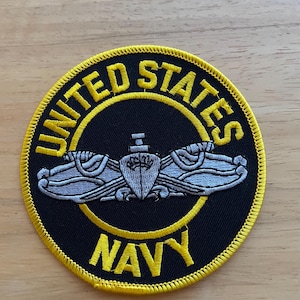 May include: A circular patch with a black background and yellow border. The words "UNITED STATES NAVY" are in yellow. A silver emblem is in the centre. The patch is likely an insignia or emblem.