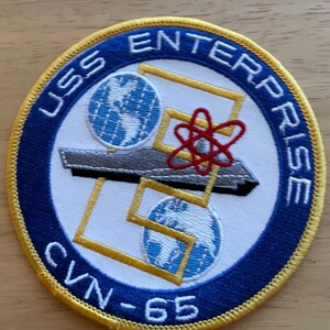 May include: Circular embroidered patch with the text "USS ENTERPRISE CVN-65". The design includes a grey aircraft carrier, two globes, and a red atom symbol, set within a blue and gold border. A nautical themed item.