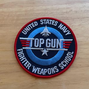 May include: Circular patch with the text "UNITED STATES NAVY" and "TOP GUN FIGHTER WEAPONS SCHOOL" in white lettering. The patch features a jet graphic, a star, and red and blue accents.