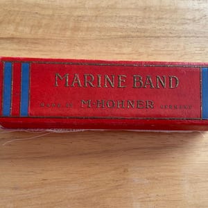 May include: A red box with the words "MARINE BAND" in gold lettering. The box has blue and gold vertical stripes on the sides. The text "MADE BY M. HOHNER GERMANY" is also visible.