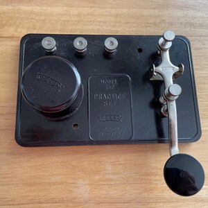 May include: A black Morse code practice set with silver keys and knobs. The set features a round black knob and a lever with a black circular end. The text "MODEL 450 PRACTICE SET" is visible.