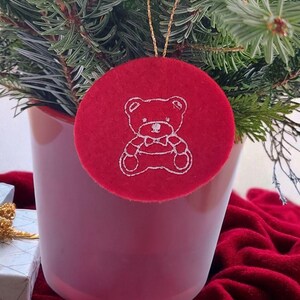 以下が含まれることがあります： 白いテディベアのデザインが刺繍された赤いフェルトのクリスマスオーナメント。 金色のチェーンで吊り下げられ、鉢植えの常緑樹の前に飾られています。 背景は赤いベルベットの布です。
