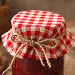 Op de afbeelding: Een pot zelfgemaakte aardbeienjam met een rood-wit geruite stoffen deksel vastgebonden met touw. Een verse aardbei ligt naast de pot op een jute doek. Een mand met aardbeien staat op de achtergrond.