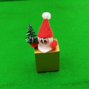 Può includere: Una piccola statuina di Natale in feltro che rappresenta Babbo Natale che tiene un albero di Natale verde. La statuina è seduta su una scatola dorata.