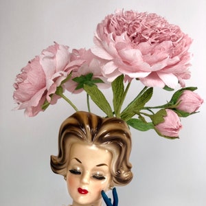 Op de afbeelding: Een vintage keramische vaas in de vorm van een vrouwenhoofd met een blauwe jurk en een blauwe handschoen. De vaas is gevuld met roze papieren bloemen.