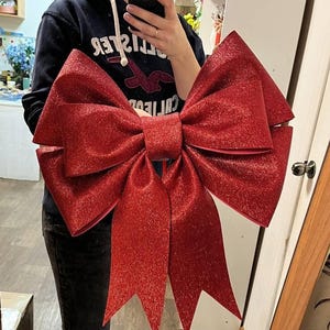 Pode incluir: Um laço decorativo grande com um acabamento de glitter vermelho vibrante. O laço apresenta múltiplas alças em camadas e duas pontas longas e afiadas que pendem para baixo. A superfície é coberta por glitter vermelho fino e brilhante.