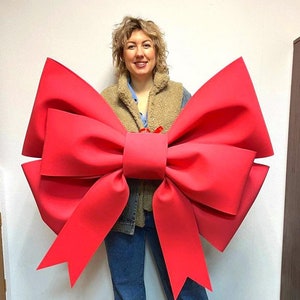 May include: A woman is wearing a large, bright red bow. The bow is made of fabric and is tied around her waist. She is standing in front of a white wall.