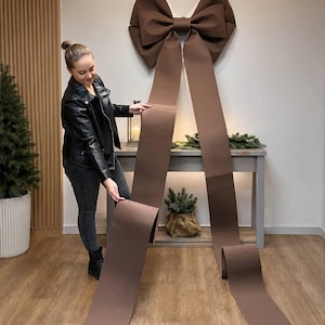 Könnte beinhalten: Eine große, matte braune Dekoschleife mit zwei langen, herabhängenden Bändern, die bis zum Boden reichen. Die Schleife ist aus strukturierten, gefalteten Schlaufen gefertigt und endet an jedem Bandende in einer klassischen V-förmigen Kerbe.