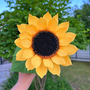 Peut inclure: Un grand tournesol en papier fait &agrave; la main, avec des p&eacute;tales jaune vif et un centre brun fonc&eacute;. La fleur est pr&eacute;sent&eacute;e sur un fond de feuillage vert et de ciel bleu. Les p&eacute;tales ont un aspect textur&eacute; et froiss&eacute;.