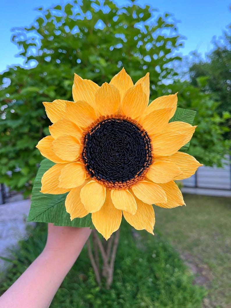 Peut inclure: Un grand tournesol en papier fait &agrave; la main, avec des p&eacute;tales jaune vif et un centre brun fonc&eacute;. La fleur est pr&eacute;sent&eacute;e sur un fond de feuillage vert et de ciel bleu. Les p&eacute;tales ont un aspect textur&eacute; et froiss&eacute;.