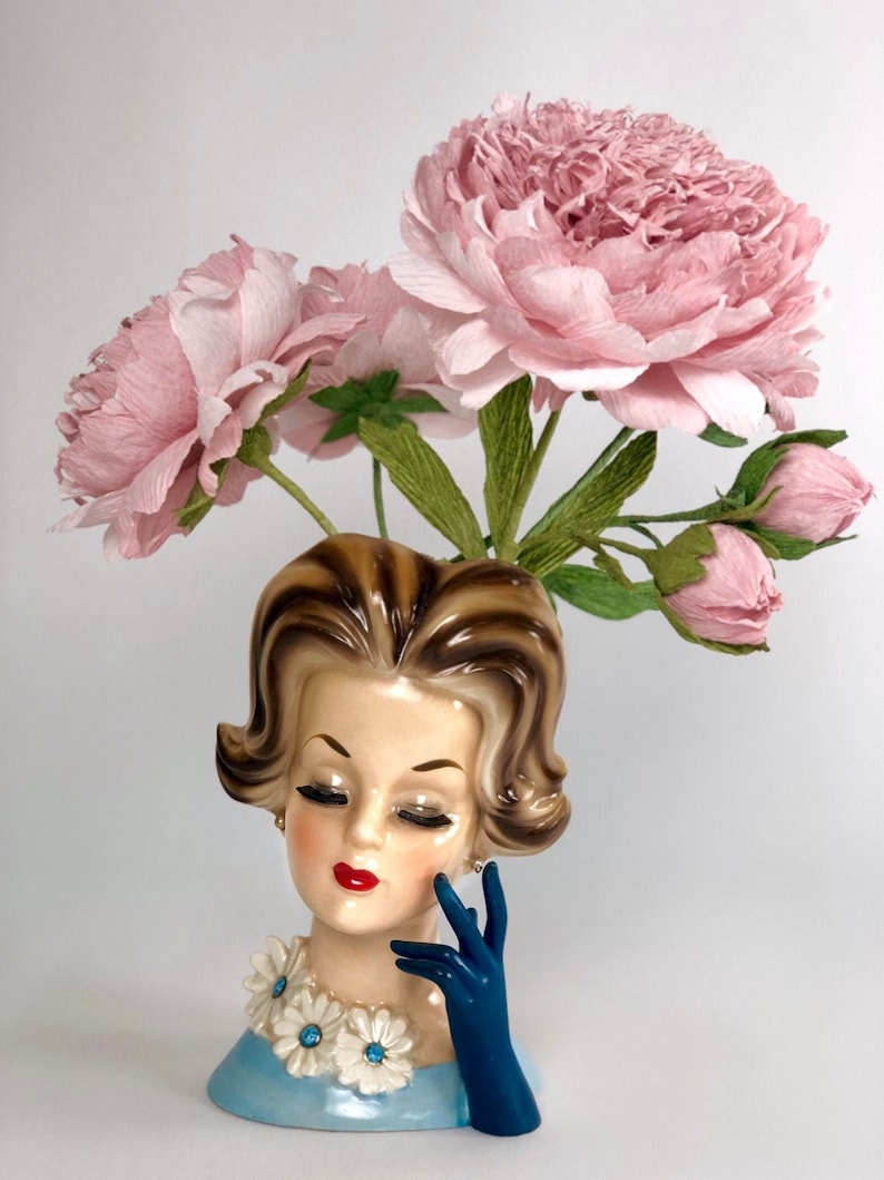 Op de afbeelding: Een vintage keramische vaas in de vorm van een vrouwenhoofd met een blauwe jurk en een blauwe handschoen. De vaas is gevuld met roze papieren bloemen.