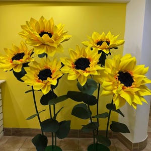 Puede incluir: Un conjunto de cinco girasoles artificiales grandes y sobredimensionados con p&eacute;talos de color amarillo brillante y centros oscuros y texturizados. Cada flor est&aacute; montada sobre un tallo verde fino y alto con hojas oscuras sobre una base circular.