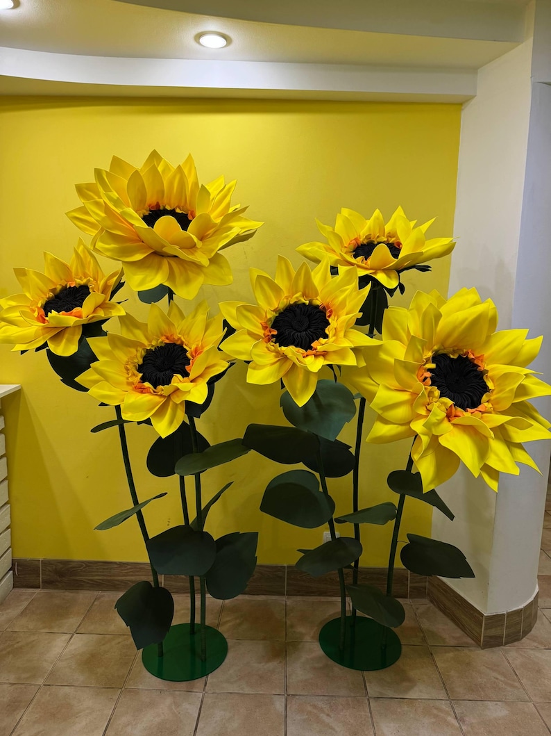 Puede incluir: Un conjunto de cinco girasoles artificiales grandes y sobredimensionados con p&eacute;talos de color amarillo brillante y centros oscuros y texturizados. Cada flor est&aacute; montada sobre un tallo verde fino y alto con hojas oscuras sobre una base circular.
