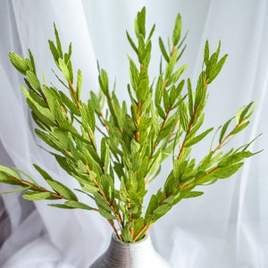 Op de afbeelding: Een boeket groene, bladerrijke takken in een zilveren vaas. De vaas heeft een getextureerd oppervlak en een ronde vorm.