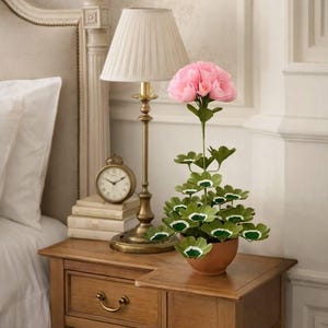 Puede incluir: Mesita de noche de madera con lámpara de latón, libros y reloj pequeño. Planta decorativa con hojas verdes y flores rosas en maceta de terracota. Decoración de dormitorio.