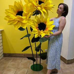 Puede incluir: Una gran escultura decorativa de suelo que presenta tres girasoles de gran tama&ntilde;o con p&eacute;talos de color amarillo brillante y centros oscuros. Las flores est&aacute;n unidas a tallos verdes altos y delgados con hojas verde oscuro, sobre una base circular verde.