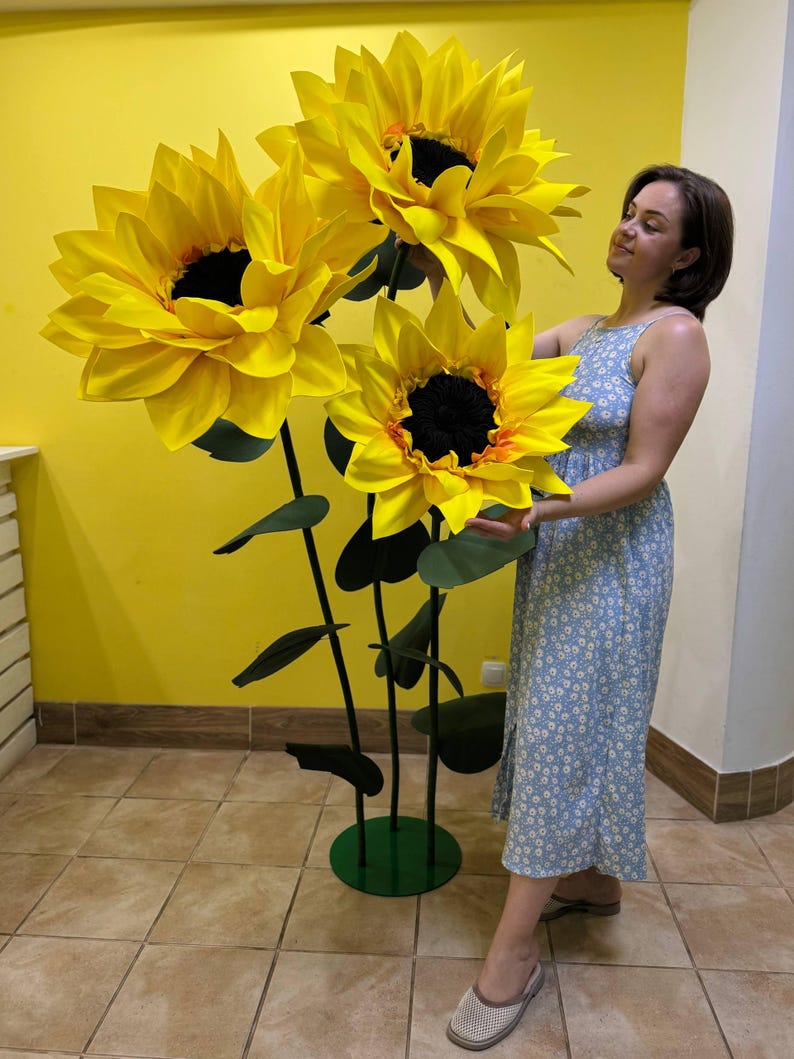 Puede incluir: Una gran escultura decorativa de suelo que presenta tres girasoles de gran tama&ntilde;o con p&eacute;talos de color amarillo brillante y centros oscuros. Las flores est&aacute;n unidas a tallos verdes altos y delgados con hojas verde oscuro, sobre una base circular verde.