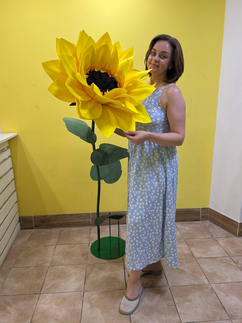 Puede incluir: Una escultura decorativa de girasol de gran tama&ntilde;o con p&eacute;talos de color amarillo brillante y un centro oscuro y texturizado. La flor est&aacute; unida a un tallo verde alto y delgado con varias hojas verdes, apoyada sobre una base circular verde.
