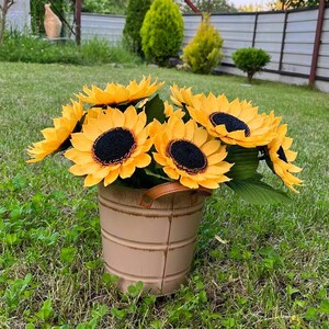 Peut inclure: Un bouquet de tournesols jaunes avec des centres noirs et des feuilles vertes dans un seau en m&eacute;tal beige avec une poign&eacute;e marron. Le seau est pos&eacute; sur une pelouse verte.