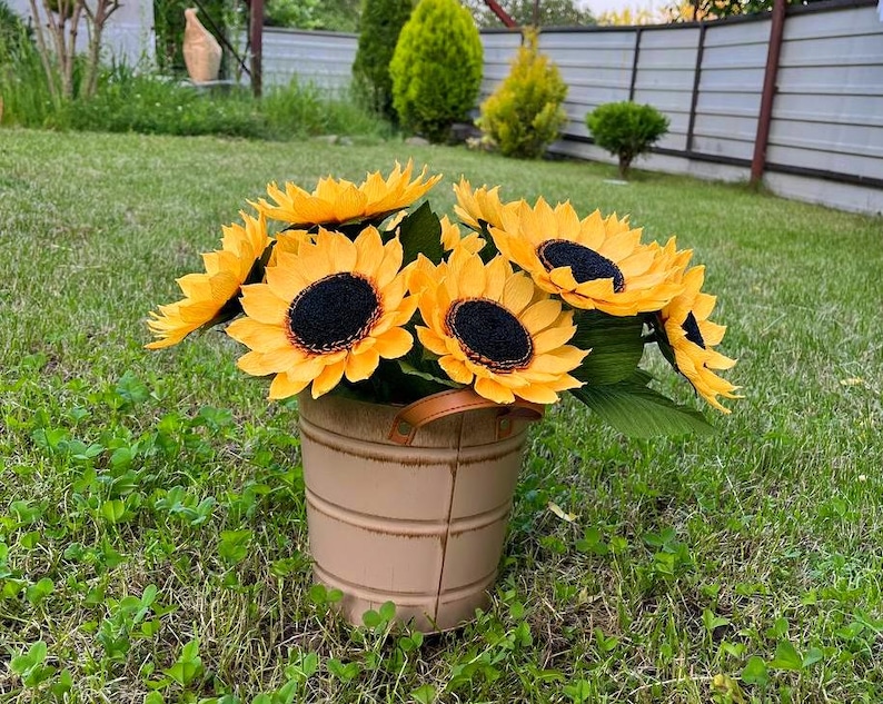 Peut inclure: Un bouquet de tournesols jaunes avec des centres noirs et des feuilles vertes dans un seau en m&eacute;tal beige avec une poign&eacute;e marron. Le seau est pos&eacute; sur une pelouse verte.