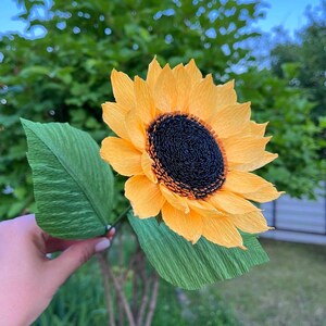 Peut inclure: Un tournesol en papier fait &agrave; la main avec des p&eacute;tales jaune vif et un centre brun fonc&eacute;. La fleur a deux grandes feuilles vertes et est tenue par une fine tige verte. L'arri&egrave;re-plan est flou, montrant le feuillage vert et un ciel bleu.