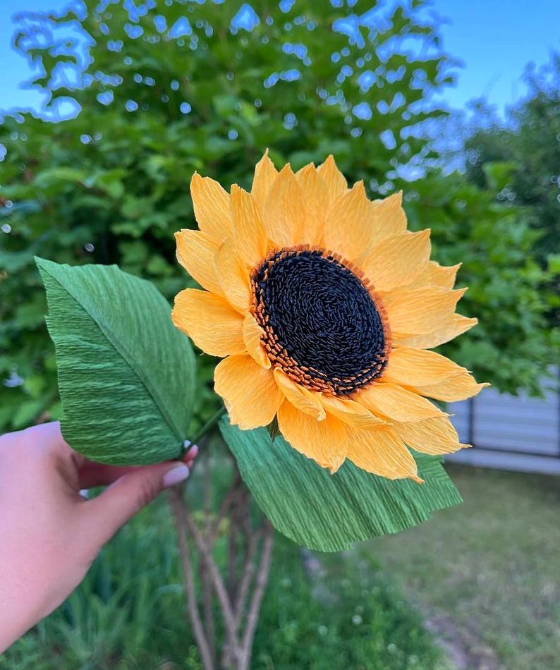Peut inclure: Un tournesol en papier fait &agrave; la main avec des p&eacute;tales jaune vif et un centre brun fonc&eacute;. La fleur a deux grandes feuilles vertes et est tenue par une fine tige verte. L'arri&egrave;re-plan est flou, montrant le feuillage vert et un ciel bleu.