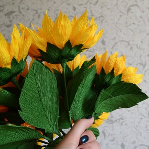 Peut inclure: Un bouquet de tournesols en papier faits &agrave; la main, avec des p&eacute;tales jaune vif et des feuilles vertes. Les fleurs sont regroup&eacute;es, mettant en valeur les d&eacute;tails de l'artisanat en papier. Le fond est un mur clair et gris &agrave; motifs.