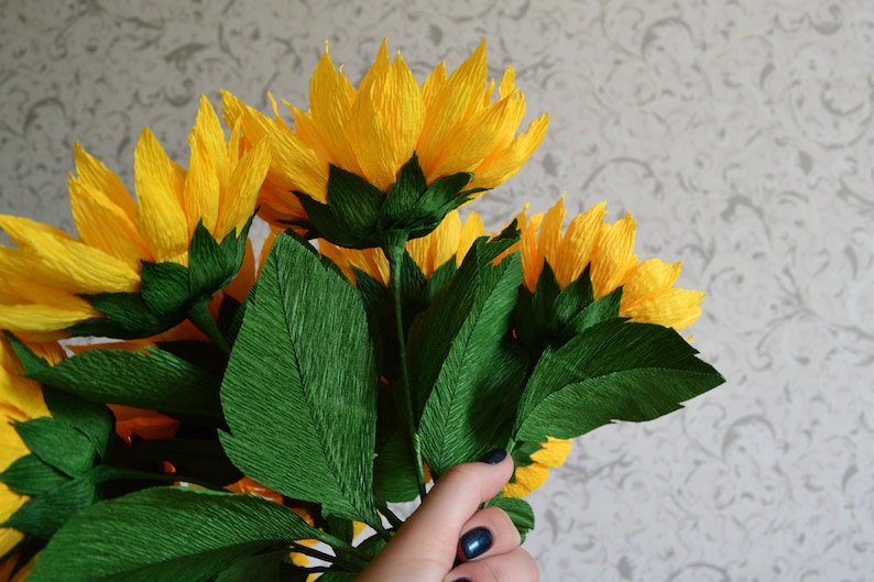 Peut inclure: Un bouquet de tournesols en papier faits &agrave; la main, avec des p&eacute;tales jaune vif et des feuilles vertes. Les fleurs sont regroup&eacute;es, mettant en valeur les d&eacute;tails de l'artisanat en papier. Le fond est un mur clair et gris &agrave; motifs.