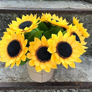 Peut inclure: Un bouquet de tournesols jaunes vifs avec des centres brun fonc&eacute; et des feuilles vertes, dispos&eacute;s dans un petit seau en m&eacute;tal blanc cass&eacute;. Les fleurs sont pos&eacute;es sur un fond de pierre vieillie.