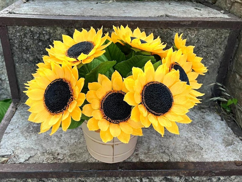 Peut inclure: Un bouquet de tournesols jaunes vifs avec des centres brun fonc&eacute; et des feuilles vertes, dispos&eacute;s dans un petit seau en m&eacute;tal blanc cass&eacute;. Les fleurs sont pos&eacute;es sur un fond de pierre vieillie.