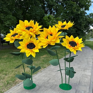 Puede incluir: Un conjunto de cinco girasoles artificiales grandes con p&eacute;talos de color amarillo brillante y centros oscuros texturizados. Cada flor est&aacute; montada sobre un tallo verde fino con varias hojas, apoyada en una base circular verde sobre un camino.