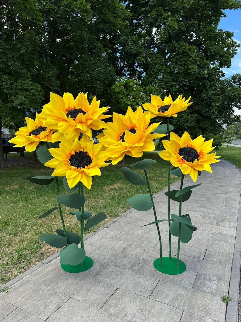 Puede incluir: Un conjunto de cinco girasoles artificiales grandes con p&eacute;talos de color amarillo brillante y centros oscuros texturizados. Cada flor est&aacute; montada sobre un tallo verde fino con varias hojas, apoyada en una base circular verde sobre un camino.