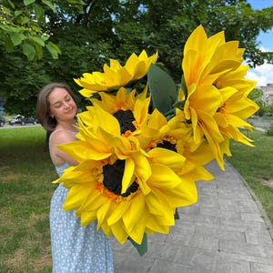 Puede incluir: Un gran ramo de girasoles artificiales de gran tama&ntilde;o con p&eacute;talos de color amarillo brillante, centros oscuros texturizados y hojas verdes. Las flores est&aacute;n construidas con p&eacute;talos en capas y se mantienen juntas en un racimo, mostrando una vibrante paleta de colores amarillos.