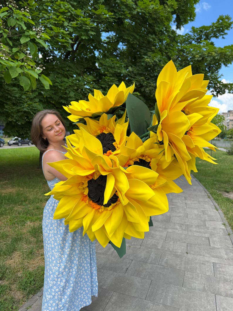 Puede incluir: Un gran ramo de girasoles artificiales de gran tama&ntilde;o con p&eacute;talos de color amarillo brillante, centros oscuros texturizados y hojas verdes. Las flores est&aacute;n construidas con p&eacute;talos en capas y se mantienen juntas en un racimo, mostrando una vibrante paleta de colores amarillos.