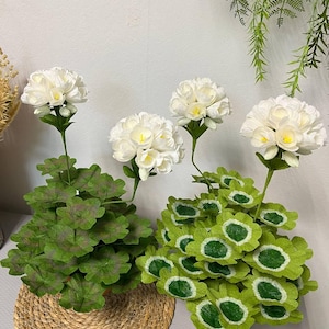May include: Artificial white geranium flowers with green leaves and stems. The flowers are arranged in two groups, one in a woven basket. The leaves have a unique shape and texture, adding visual interest to the arrangement.