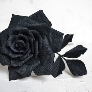 Può includere: Una singola rosa di carta nera con stelo e foglie. La rosa è fatta di carta stropicciata e ha un aspetto realistico.