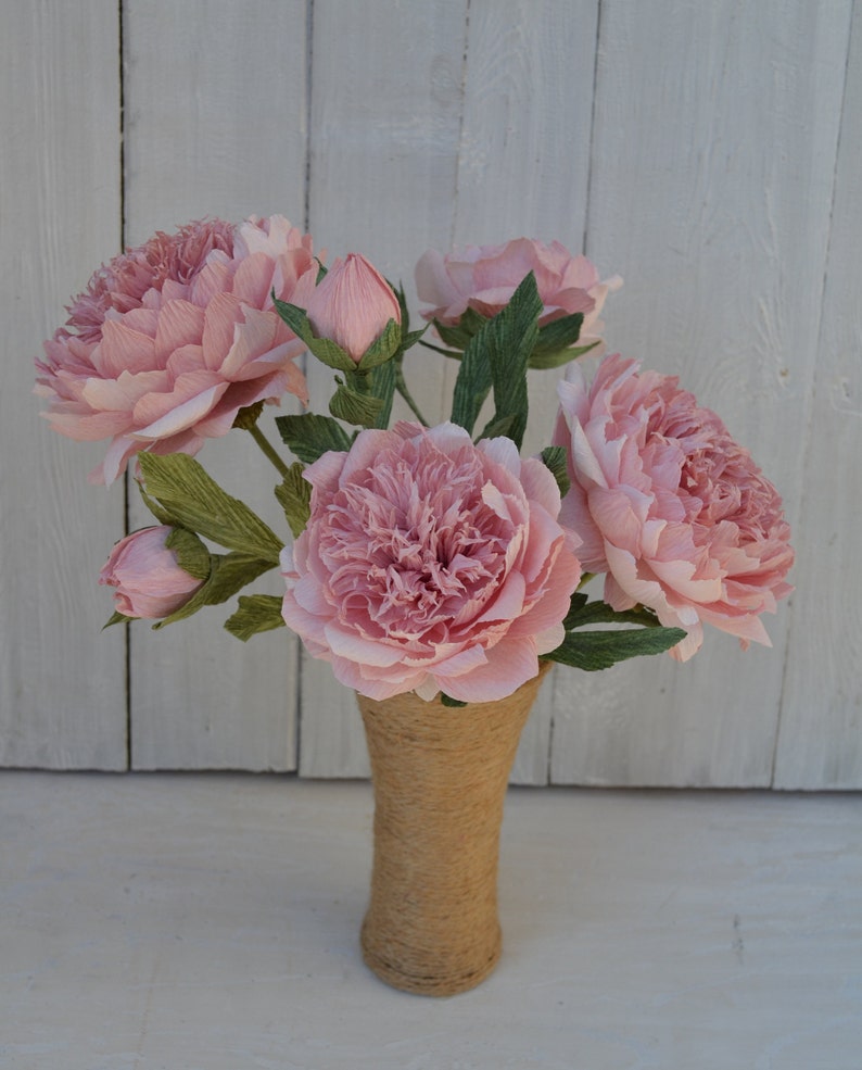 Op de afbeelding: Een boeket roze papieren pioenen in een jute vaas. De pioenen zijn gerangschikt in een vaas met een rustieke, natuurlijke uitstraling.