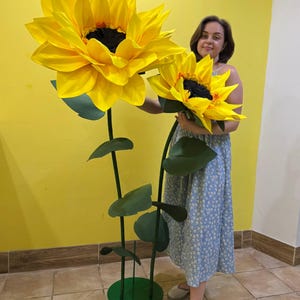 Puede incluir: Dos girasoles artificiales de gran tama&ntilde;o con p&eacute;talos de color amarillo brillante y centros de color marr&oacute;n oscuro. Cada flor est&aacute; unida a un tallo verde alto y delgado con varias hojas verdes. Los tallos se apoyan en una base circular verde.