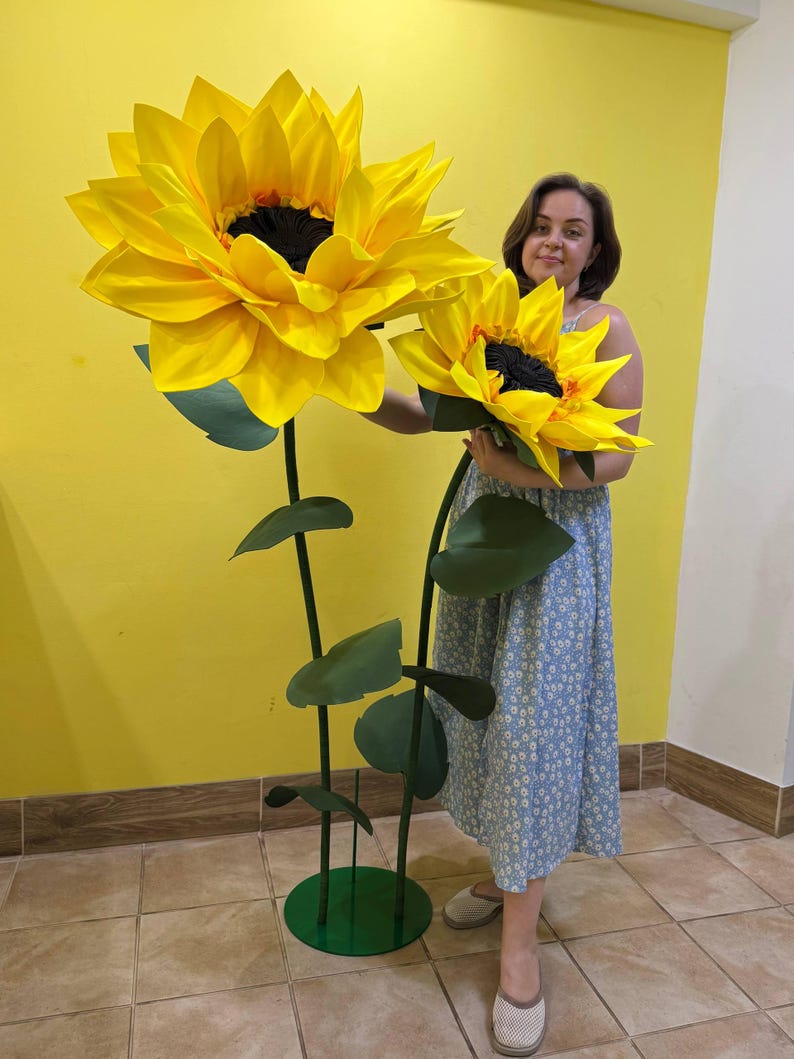 Puede incluir: Dos girasoles artificiales de gran tama&ntilde;o con p&eacute;talos de color amarillo brillante y centros de color marr&oacute;n oscuro. Cada flor est&aacute; unida a un tallo verde alto y delgado con varias hojas verdes. Los tallos se apoyan en una base circular verde.