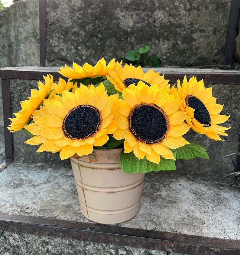 Peut inclure: Un bouquet de tournesols jaune vif avec des centres noirs et des feuilles vertes, dans un seau en m&eacute;tal beige. Les tournesols sont dispos&eacute;s pour cr&eacute;er une pr&eacute;sentation compl&egrave;te et vibrante, id&eacute;ale pour la d&eacute;coration int&eacute;rieure ou les cadeaux.