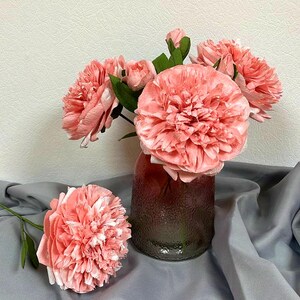 Op de afbeelding: Een boeket roze papieren bloemen in een glazen vaas met een getextureerd oppervlak. De vaas staat op een grijs stoffen oppervlak.