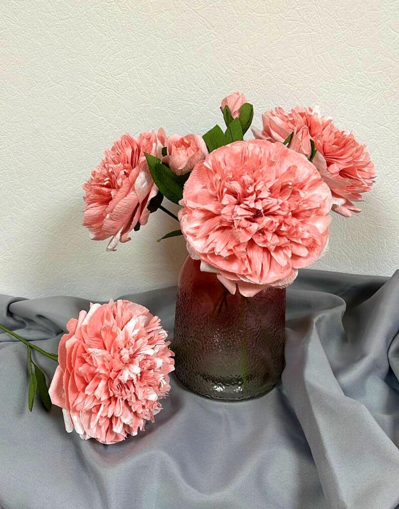 Op de afbeelding: Een boeket roze papieren bloemen in een glazen vaas met een getextureerd oppervlak. De vaas staat op een grijs stoffen oppervlak.